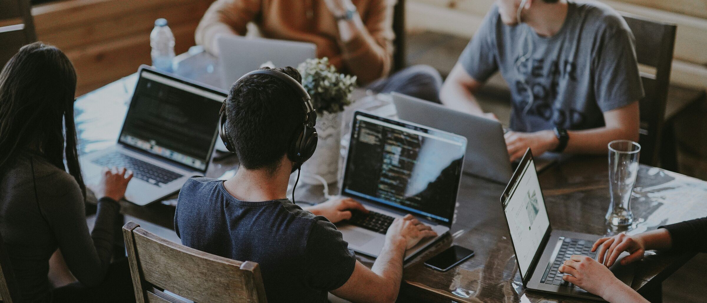 Five people from different companies working at one wooden table, each behind a laptop, in conversation about what is on the screens.