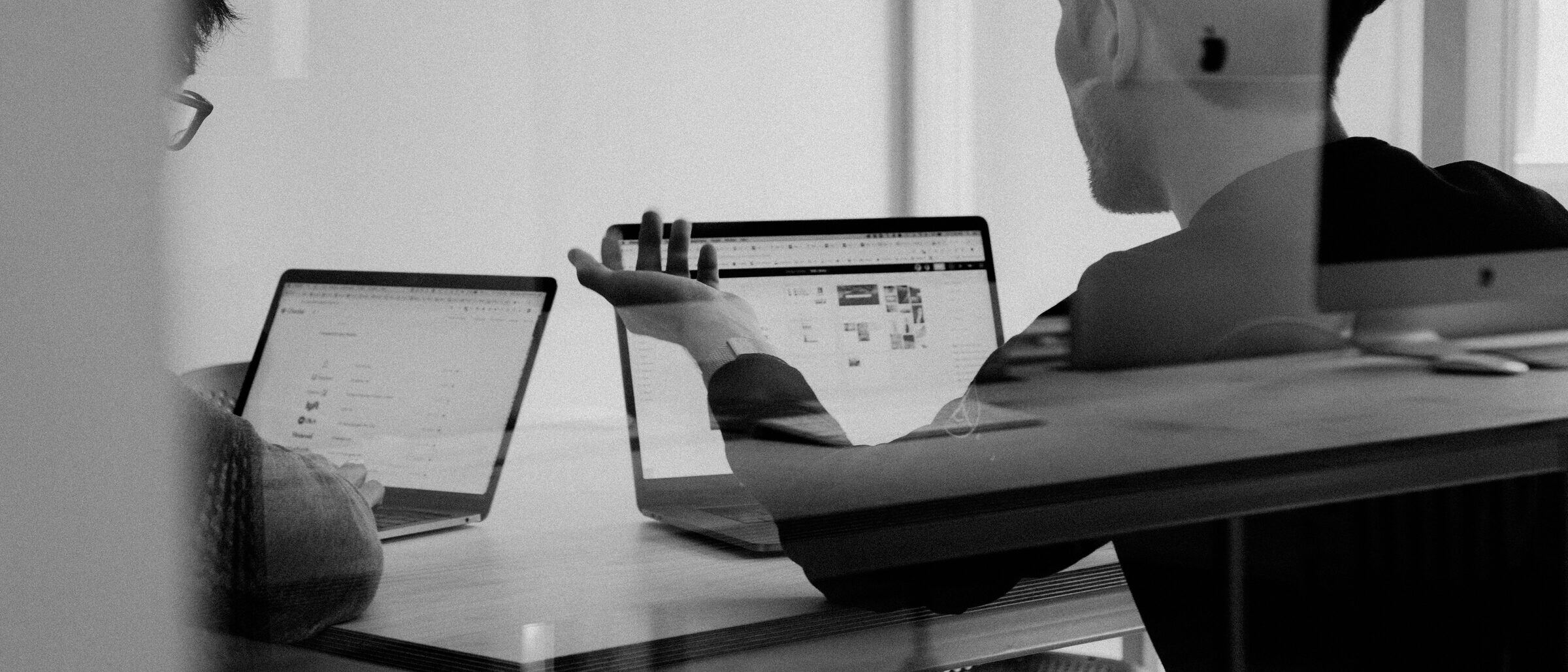 Two people working at their laptops at a shared table, seen through a glass partition.