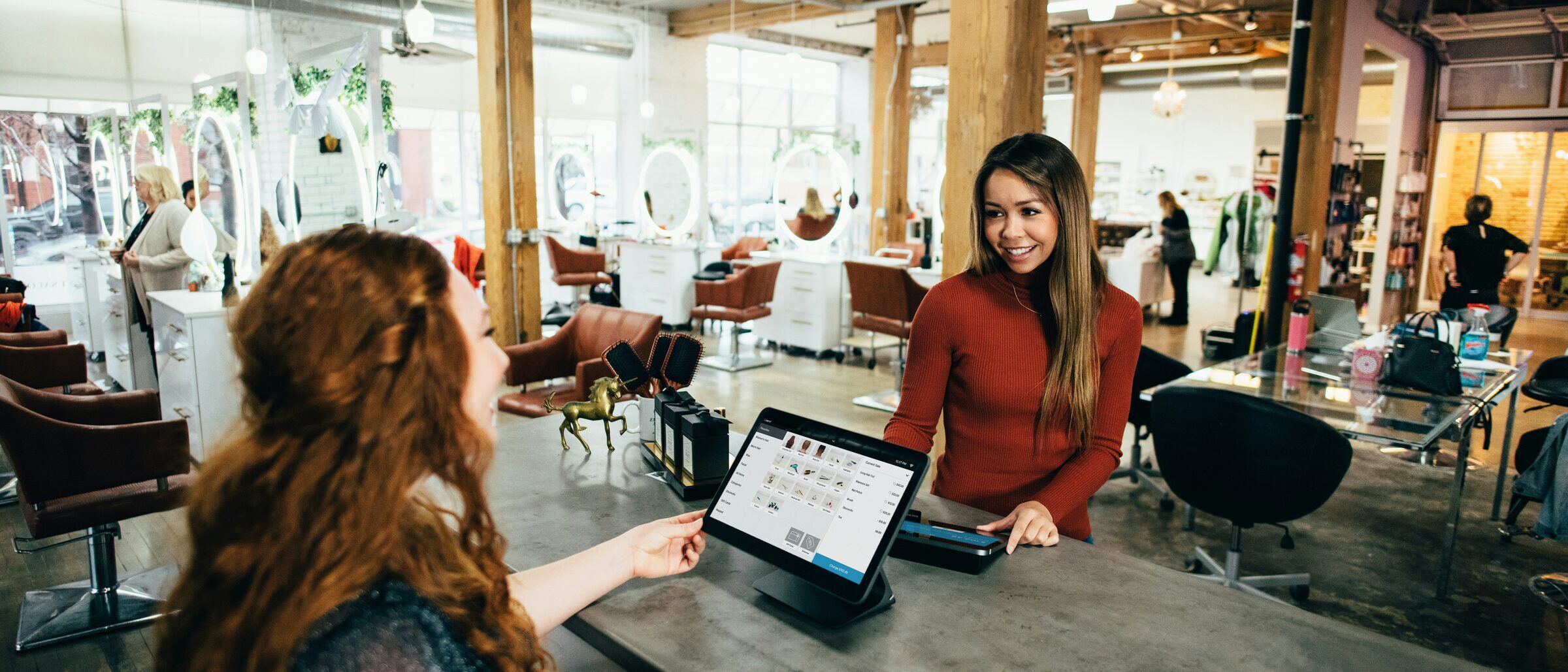 An employee helping a customer at a shop counter, with a tablet checkout between them.